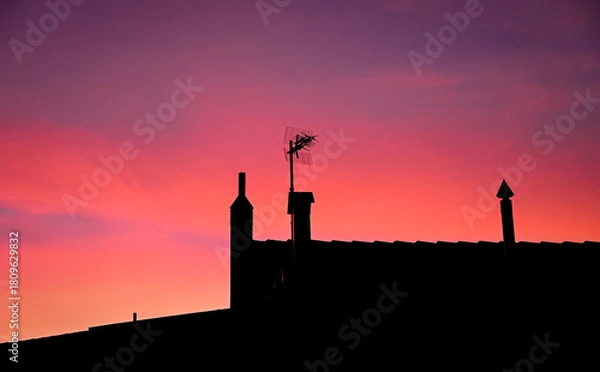 Obraz Silhouette of a rooftop with chimneys and a TV antenna against a vivid red and purple sunset sky, creating a dramatic and peaceful evening atmosphere