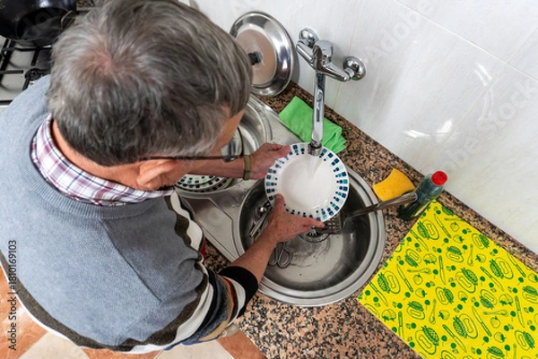 Fototapeta A senior man washes a ceramic plate under running water at the kitchen sink, performing a routine household cleaning task from an overhead view.