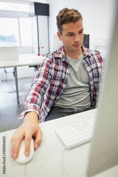 Obraz Man in 20s sitting in workspace using computer with keyboard and wired mouse, wearing plaid shirt