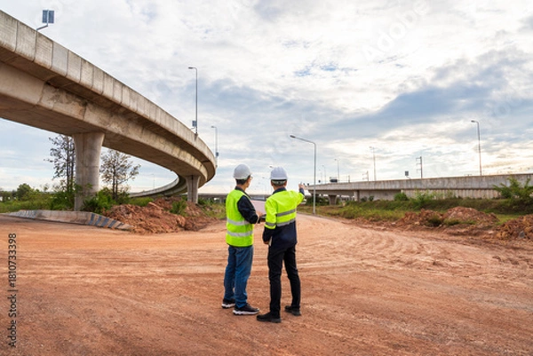 Fototapeta Two construction engineers stand on a vast dirt road, inspecting the site under a sweeping curved concrete highway overpass.