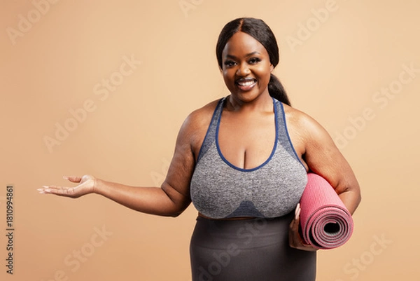 Obraz Smiling plus size model holding yoga amt showing with hand, copy space isolated on beige background
