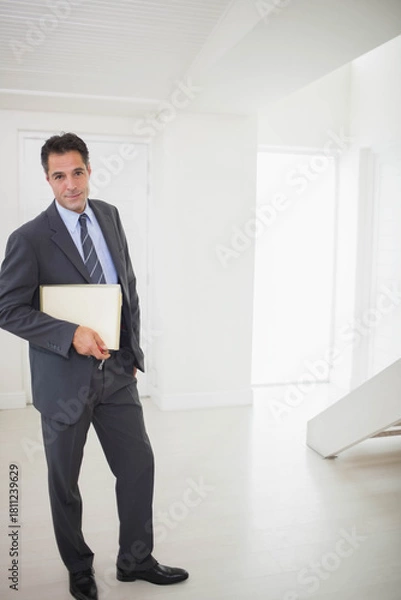 Fototapeta Mature man standing left in bright entryway holding beige folder keys wearing suit, copy space
