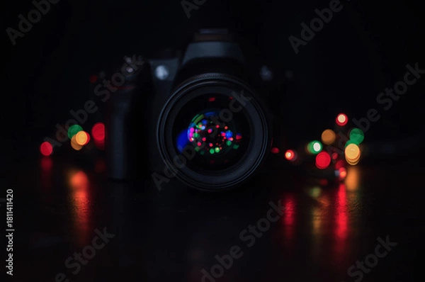 Fototapeta Close-up DSLR camera on dark background with festive bokeh lights reflected in the lens. Creative photography concept, low key, holiday ambiance.
