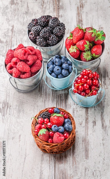 Fototapeta Berries colorful closeup assorted mix in cans and small knitted basket. Raspberry, blueberry, red currant, strawberry on white rustic wooden table in studio.