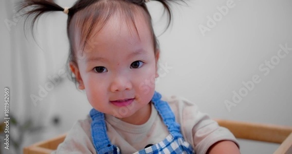 Fototapeta Cute Asian toddler smiling and looking at camera, wearing a blue checkered apron, creating a joyful and playful atmosphere.
