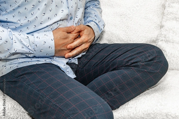 Fototapeta A man in elegant trousers and a long blue shirt sits on a white sofa, holding his aching stomach, hands on his stomach.