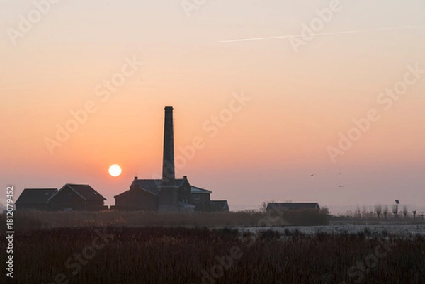 Fototapeta A sunrise over a field with a large brick building in the background. The sky is orange and the sun is rising.