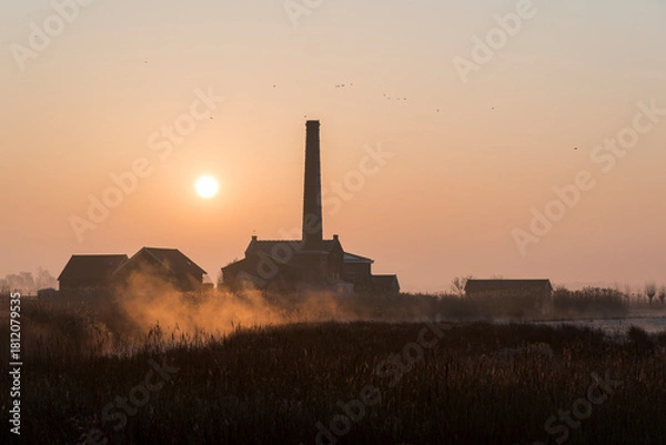 Fototapeta A sunrise over a field with a large brick building in the background. The sky is orange and the sun is rising.