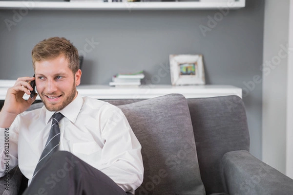 Obraz Adult man sitting on sofa in living room holding smartphone and wearing striped tie, copy space