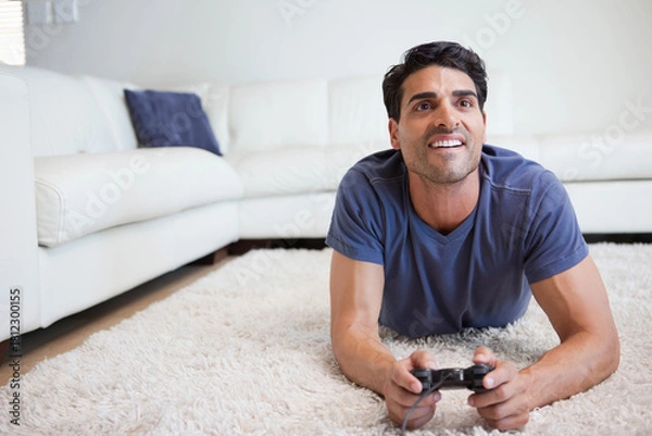 Fototapeta Man lying on shag rug holding wired game controller wearing blue tee in living room