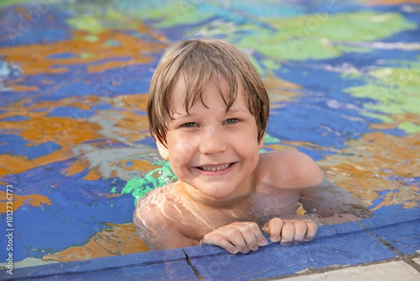 Fototapeta Smiling boy enjoying time in an outdoor swimming pool, holding onto the pool edge on a sunny day. Summer vacation and fun water activities for kids.