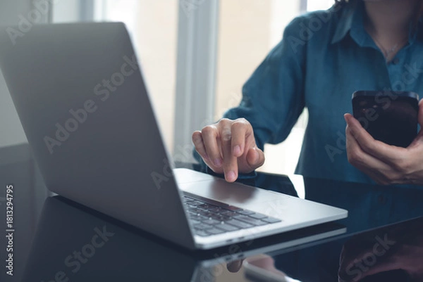 Obraz Businesswoman working on laptop computer and using mobile phone at office. Asian woman using smartphone searching the information