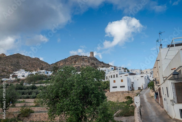 Fototapeta The Atalaya de Nijar, a historic watchtower in Almeria, Spain, stands atop a rocky hill. The structure features a cylindrical design, surrounded by sparse greenery under a bright, partly cloudy sky