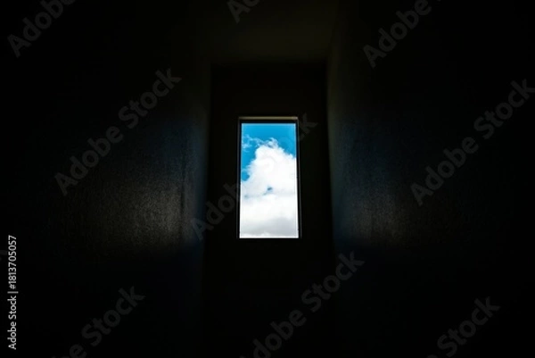 Fototapeta View of bright blue sky with white clouds through a narrow rectangular window in a dark interior space creating a contrast of light and shadow