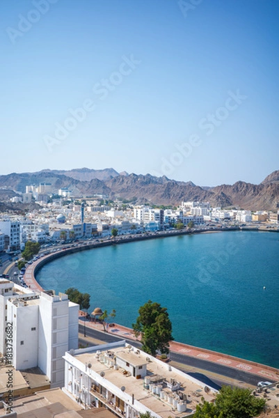Fototapeta High-resolution vertical photo of the Muttrah Corniche in Muscat, Oman, featuring a sweeping curved waterfront lined with white Arabian buildings, turquoise blue water, and rugged rocky mountains