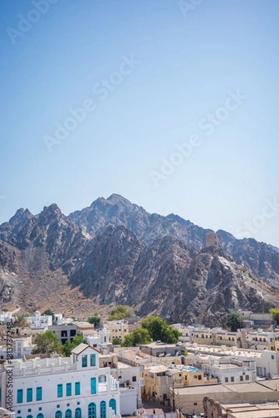 Fototapeta High-resolution vertical photo of Old Muscat, Oman, featuring rugged rocky mountains rising behind traditional white and beige Middle Eastern buildings. A historic watchtower