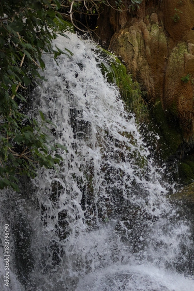 Obraz A powerful waterfall crashes over moss-covered rock formations, surrounded by lush greenery, capturing the raw motion and texture of flowing water in nature.