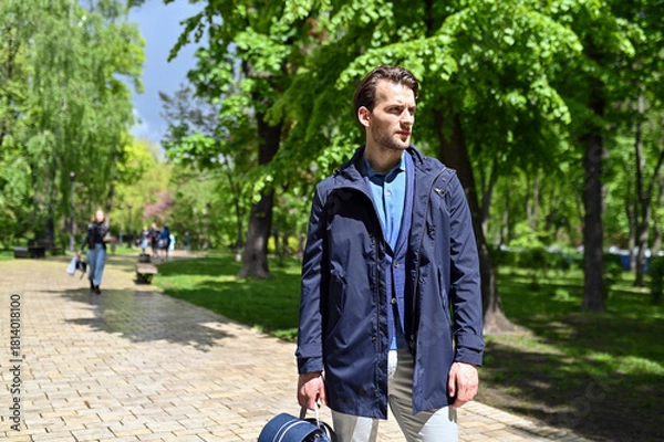Obraz A stylish man walks through a sunlit park wearing a navy jacket and light trousers. Fresh greenery, soft shadows and his relaxed stride create a calm lifestyle atmosphere.