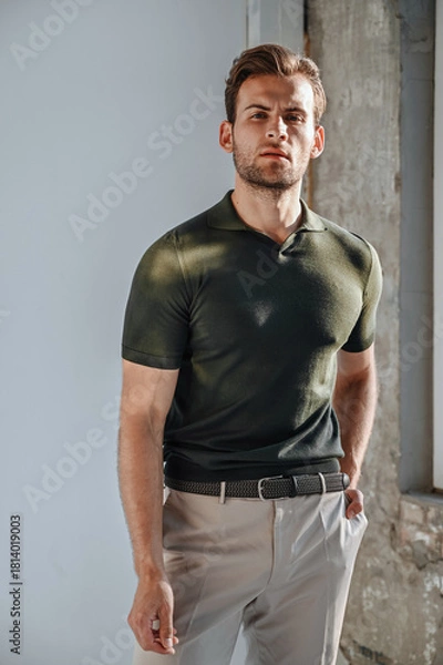 Obraz A man in a green T-shirt poses against a concrete wall, standing casually with one knee bent. The raw texture of the background adds a bold, contemporary fashion edge.
