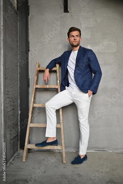 Obraz A man in a navy blazer stands by a wooden ladder in a textured concrete room, blending modern fashion with rustic industrial style.