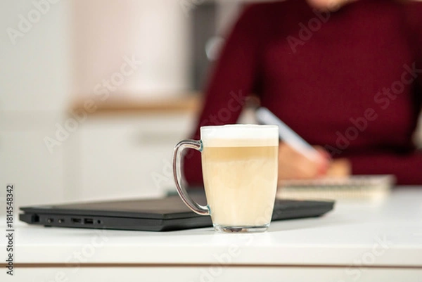 Obraz Fresh cappuccino in a glass mug on a white table with a woman working on a laptop in the blurred background. Ideal for remote work and cafe concepts.