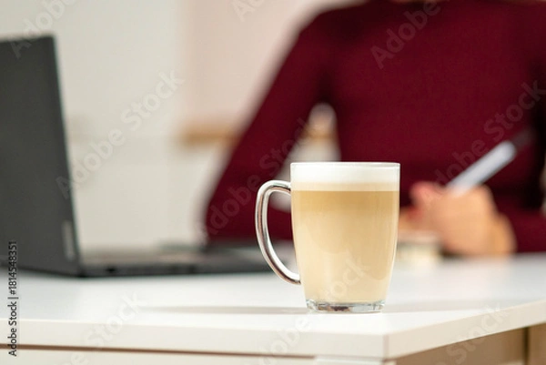 Obraz Glass mug of latte coffee in the foreground with a laptop and a woman writing in the background. Cozy remote work and studying atmosphere.