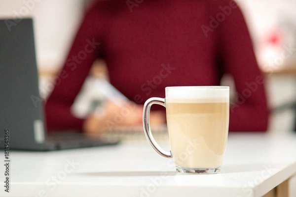 Obraz Freshly made layered cappuccino on a white desk. In the background, a woman in a red sweater is studying or working. Concept of focus and comfort.