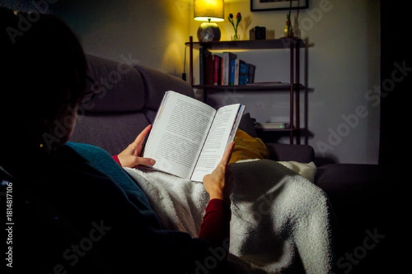 Obraz A woman wrapped in a blanket reads a book on the sofa in a dim living room, illuminated only by the warm glow of a floor lamp.