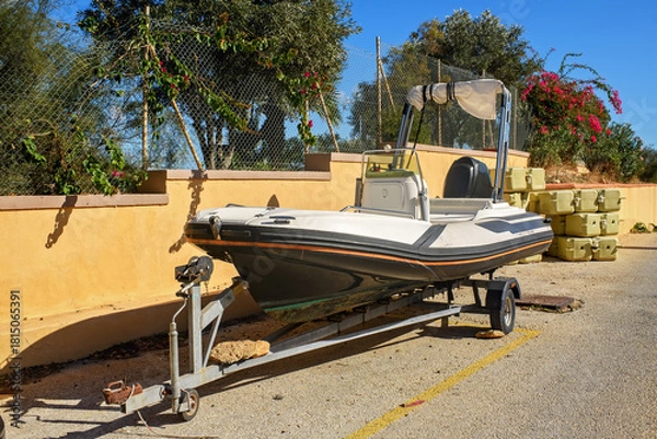 Obraz A road trailer with an inflatable motorboat is parked on the street against a wall, with flowering plants and modular pontoon blocks in the background.