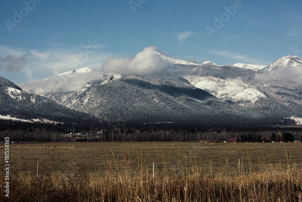 Fototapeta Snow covered mountain scene with bright blue sky over a grassy valley floor