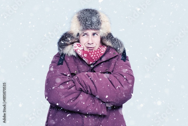 Obraz Man in red winter jacket and fur trapper hat shivers in falling snow, hugging himself for warmth against a cold, pale backdrop. Seasonal cold weather, freezing outdoor concept, harsh winter