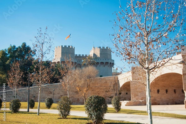 Fototapeta A view from Turia park gardens to  Serranos Gate with a flag, famous Valencian Towers, part of the ancient city wall, and a roman bridge at the foreground on autumn winter sunny day.