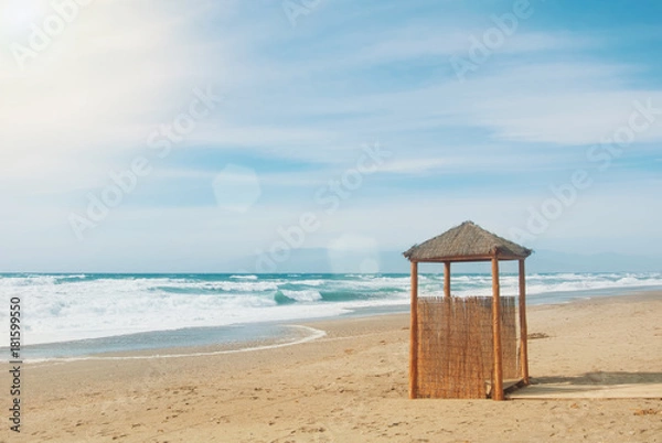 Fototapeta Wild abandoned unexplored beach with a lonely wooden wicker construction, bright blue turquoise sea ocean waves, summer paradise exotic water landscape with mountains and clouds on horizon.