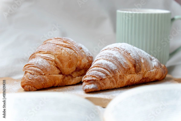 Obraz Freshly baked croissants with powdered sugar and a green coffee mug, set against a cozy bed background.
