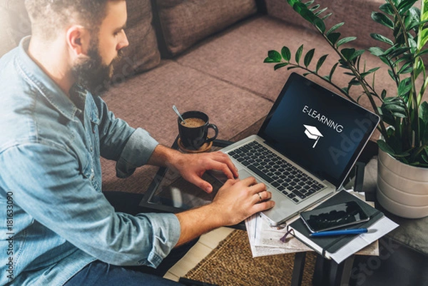 Fototapeta Młody brodaty mężczyzna hipster, przedsiębiorca siedzi na kanapie przy stoliku do kawy, korzysta z laptopa z napisem - e-learning na ekranie. Na stole notatnik, filiżanka kawy. Marketing online, edukacja, e-learning. Startup.