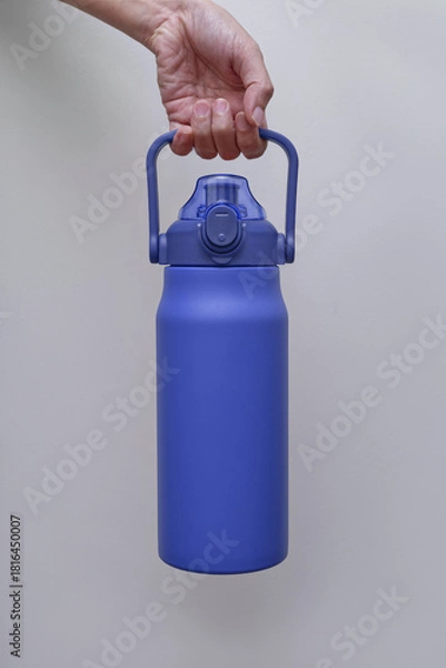 Fototapeta A hand holds a large, textured blue insulated water bottle with a convenient handle and straw lid. The bottle is presented against a neutral light grey background.