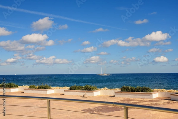 Obraz Blue summer sky with scattered white clouds over the Mediterranean Sea, featuring a sailboat and a concrete seaside promenade with potted green bushes.