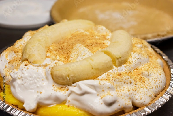 Fototapeta The finished banana cream pie, topped with fluffy whipped cream, graham cracker crumbs, and two whole peeled bananas for a perfect, decorative presentation.