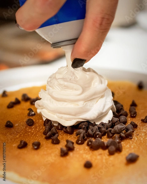 Fototapeta A chef adds a swirl of whipped cream from a can onto a fluffy pancake sprinkled with mini chocolate chips, creating a sweet breakfast.