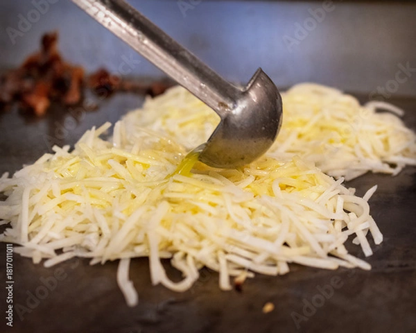 Fototapeta A chef uses a ladle to pour melted butter over shredded hash browns cooking on a hot flat-top griddle, preparing a savory breakfast.