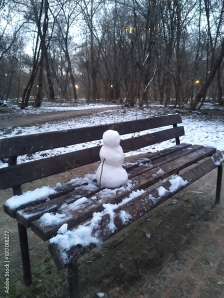 Obraz Small Snowman Sitting Alone on a Park Bench in an Early Winter Landscape with Bare Trees at Dusk_1