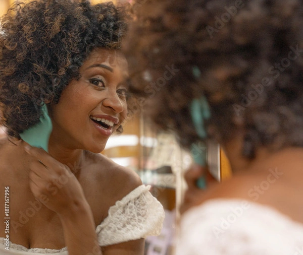 Fototapeta Elegant young woman with curly hair, looking in the mirror and smiling. She is trying on a gold necklace and a light blouse, evaluating her appearance.
