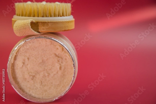 Fototapeta Exfoliating scrub jar with wooden brush on a bright red background, showcasing natural skincare products for beauty routines and self-care practices for wellness lovers