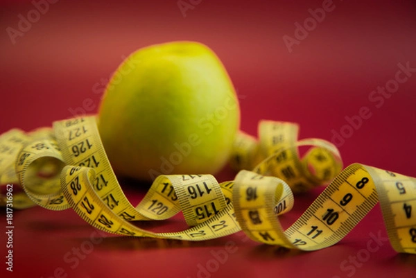 Fototapeta Fresh green apple on a striking red backdrop, entwined with a yellow measuring tape, representing health, nutrition, and the significance of mindful eating habits