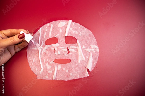 Obraz Hand of a woman applying serum to a facial mask against a bold red backdrop, highlighting skincare rituals and the importance of self-care in beauty routines
