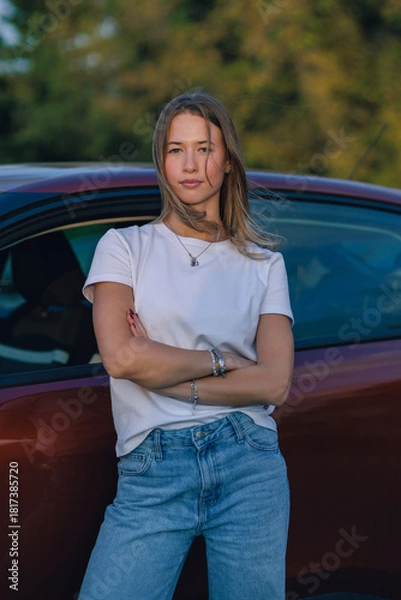 Obraz Young woman stands confidently next to her car in the evening light at a park