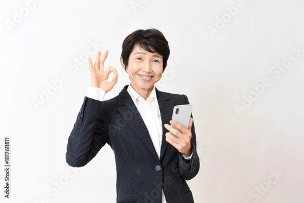 Obraz スマホを持ちながらOKサインをするスーツ姿のアジア人のミドル女性