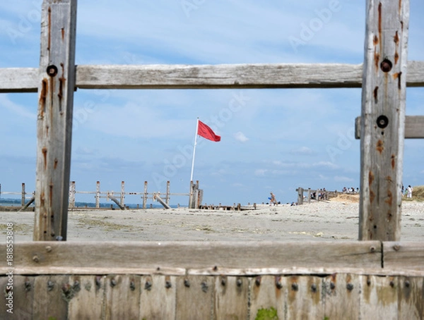 Fototapeta Red Warning Flag Flying on the Beach Framed by Weathered Wooden Groynes and View of Distant Coastal Defenses