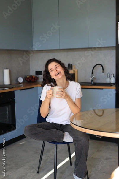 Obraz Smiling woman sitting in the kitchen with a cup of coffee, enjoying a relaxed and cheerful moment in a cozy home environment