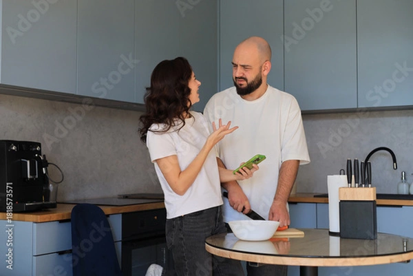 Obraz Couple having a playful discussion while preparing food in the kitchen, gesturing and interacting during a casual cooking moment
